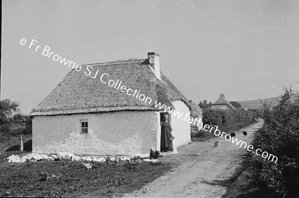 WOMAN AT DOOR OF WHITEWASHED COTTAGE